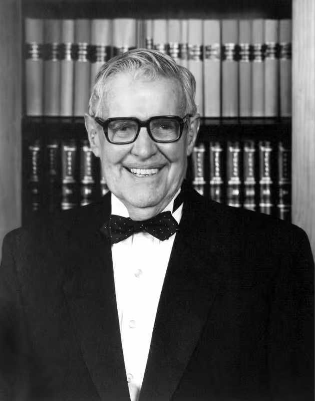 Smiling elderly man wearing glasses, a tuxedo, and a bow tie, standing in front of a bookshelf.