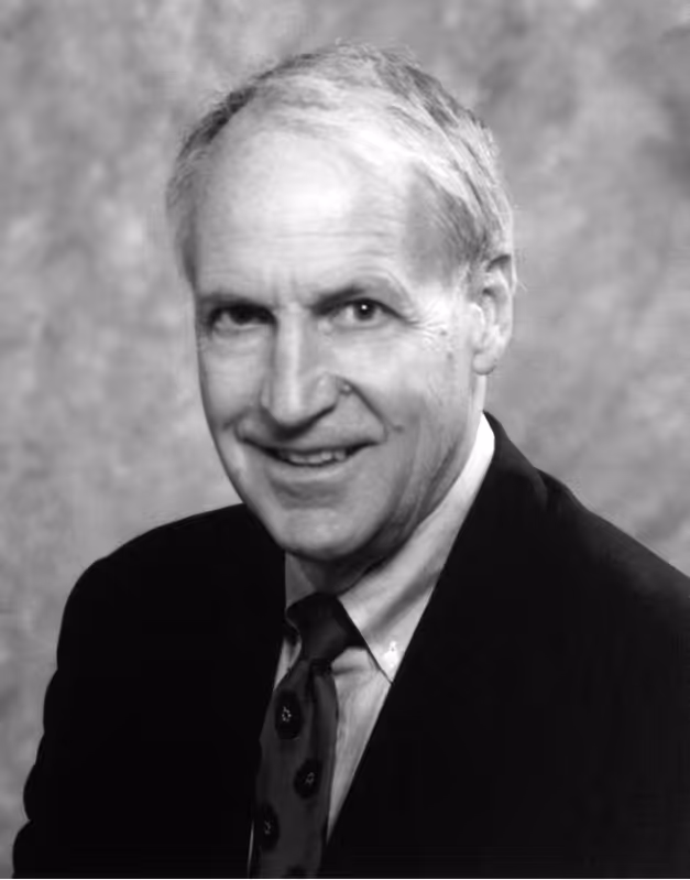 Black and white portrait of an older man smiling, wearing a suit, shirt, and patterned tie with a blurred background.