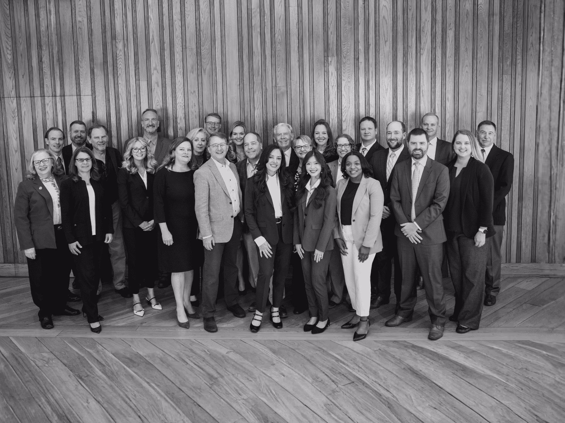 Group portrait of twenty-seven professionally dressed individuals standing indoors against wooden paneling.