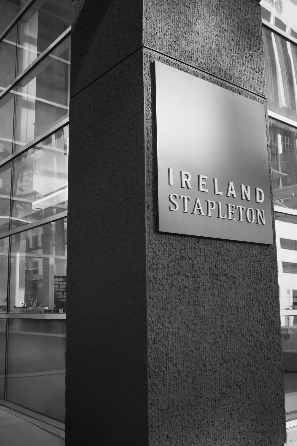 Metal plaque on a building corner reading 'IRELAND STAPLETON' next to large glass windows.