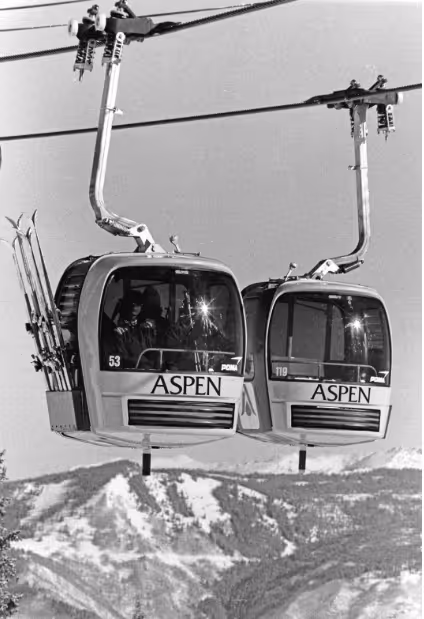 Two Aspen ski gondolas suspended on cables above snowy mountain slopes, one carrying skis.