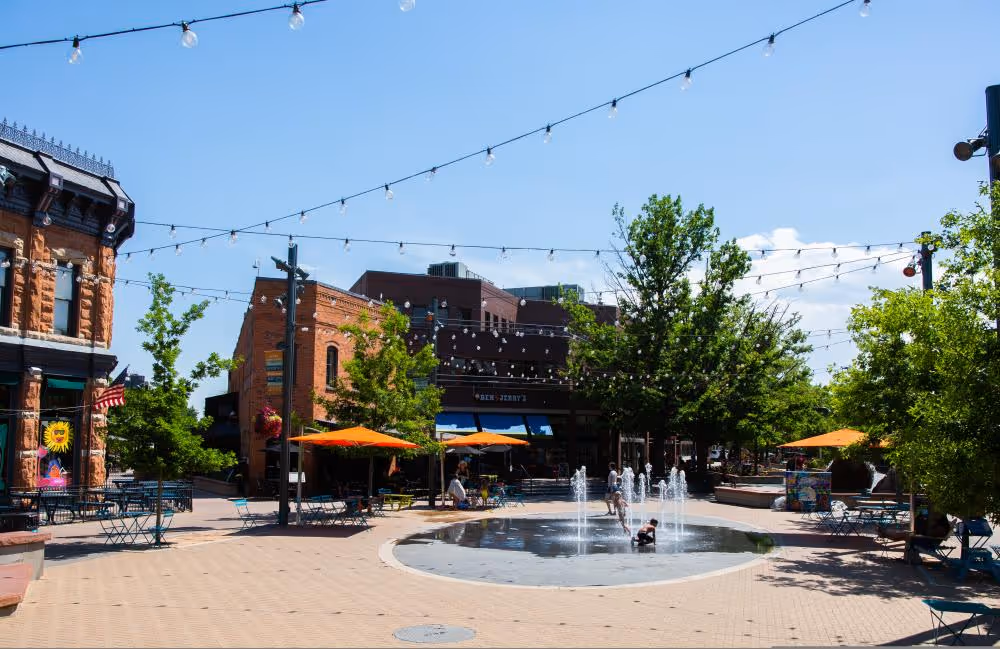 Outdoor plaza with children playing in a circular water fountain, surrounded by benches, tables with orange umbrellas, trees, and brick buildings under a clear blue sky.