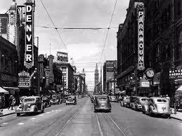 Black and white street view of vintage cars parked along buildings with Denver and Paramount signs in an urban setting.