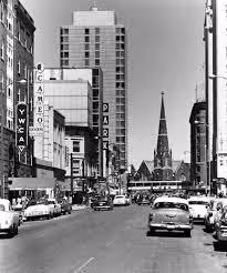 Black and white photo of a city street with vintage cars, tall buildings, and a church steeple in the background.