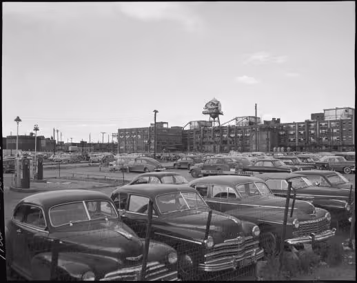Black and white photograph of a parking lot filled with vintage cars in front of industrial buildings with a large water tower in the background.