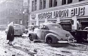 Black and white photo of a muddy street outside Denver Union Bus station with vintage cars and a woman walking with an umbrella.