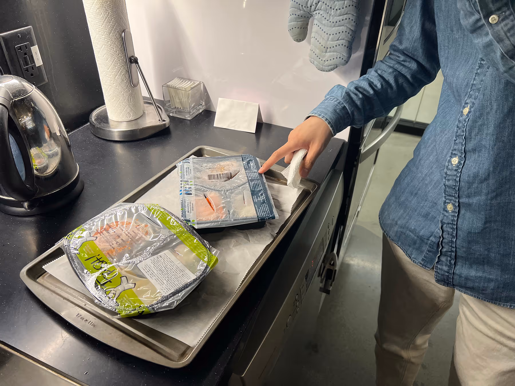 A person pointing at a prepackaged meal that has just come out of the oven. The meal container is next to another prepackaged meal container that has also been cooked, and both sit on a sheet pan to protect them while baking.
