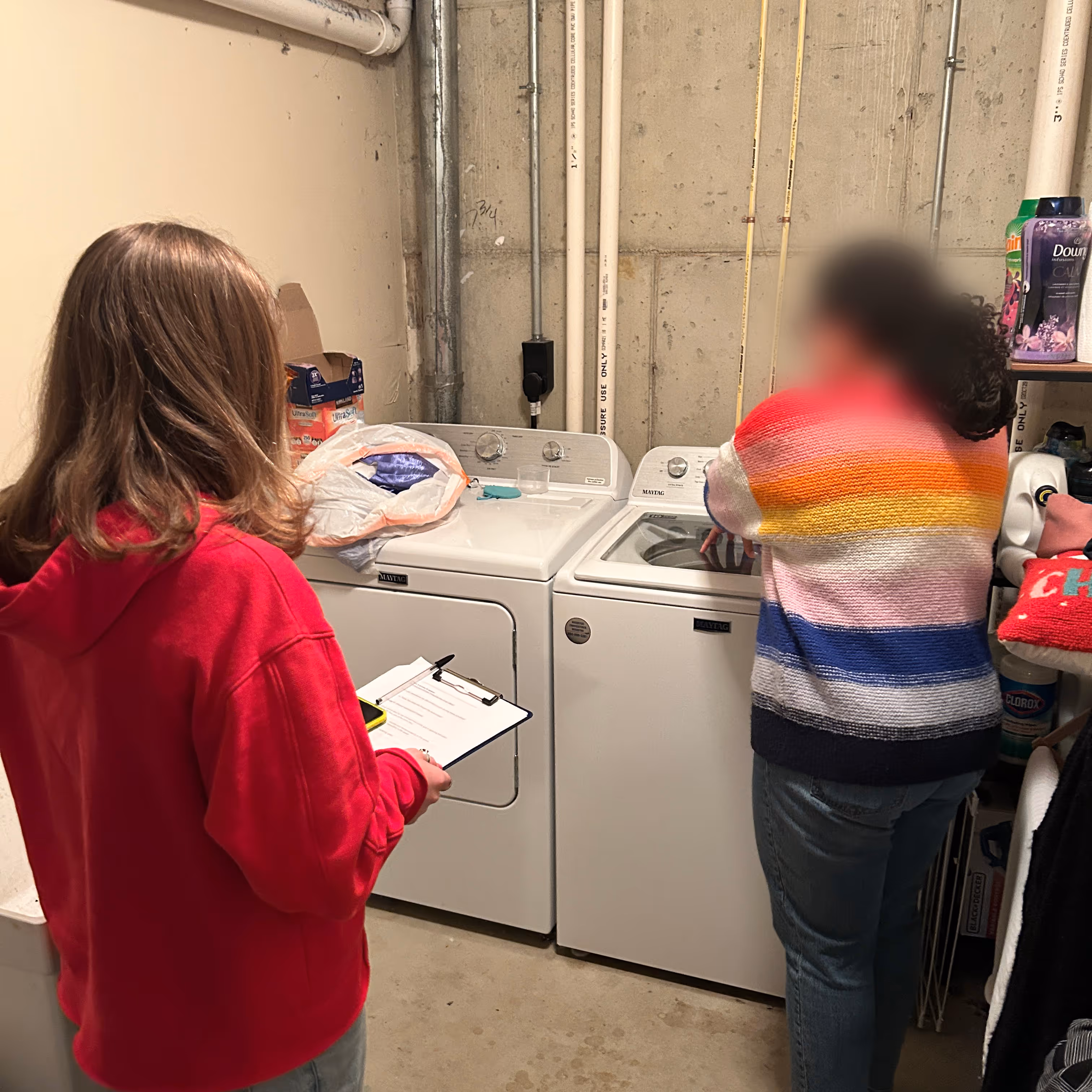 A female researcher in a red sweater observes a research participant in a brightly colored striped sweater describing her laundry process in her laundry room
