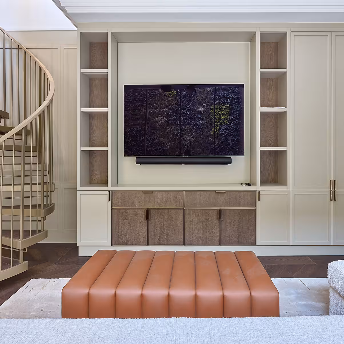 Room with a gold spiral staircase to the left, a full height unit in the centre which contains cupboards, a TV and sound bar. Directly in front there’s a burnt orange leather footstool.