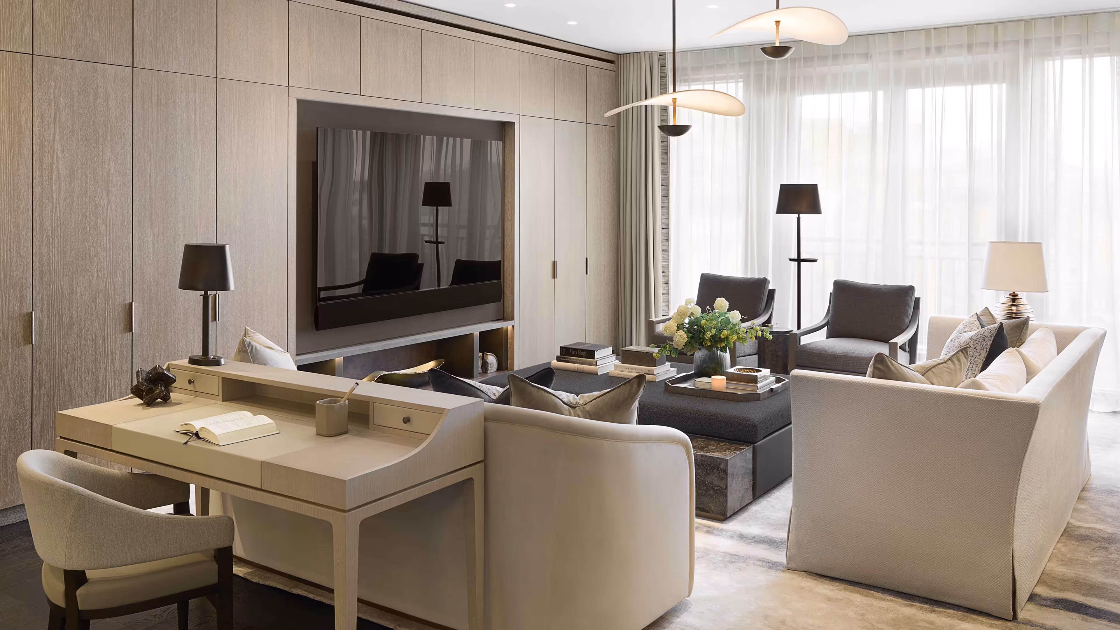 Living room with a full height wall with built in cupboards and flat screen TV to the left. In front of the TV there’s two cream sofas and two grey sitting chairs. The far wall has full height and width windows covered by light blinds.