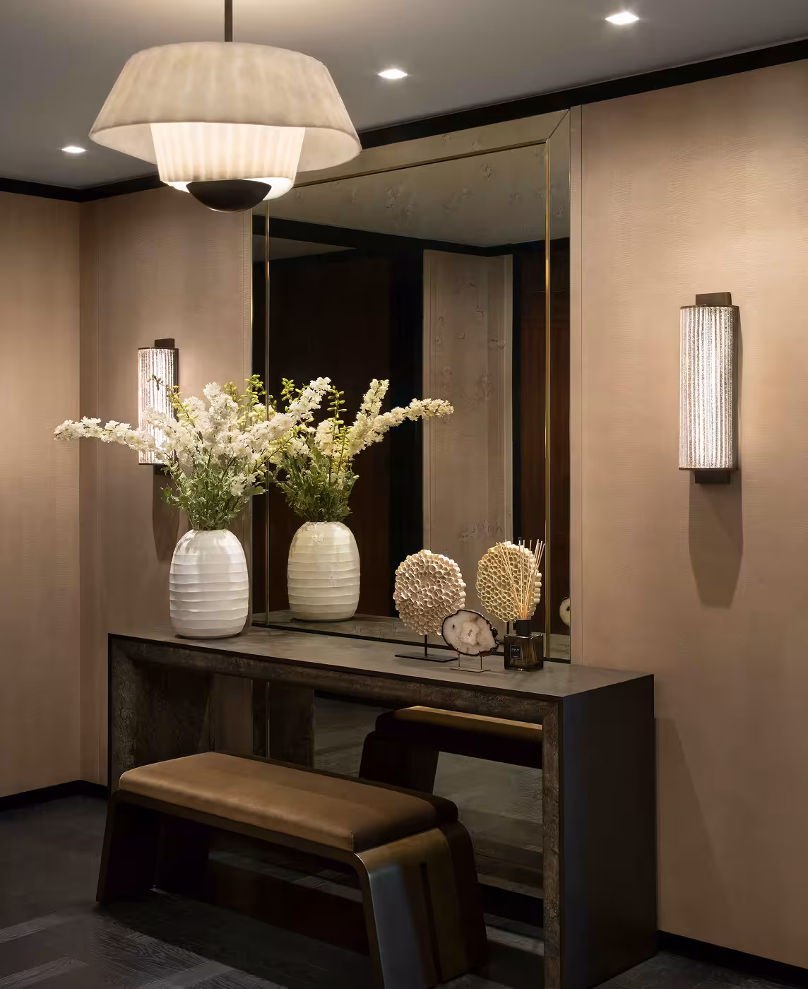 Entrance area with a mirror, sat behind a dark wood table and stool. Either side of the mirror there are two art deco style, wall mounted lights and dark beige coloured walls.