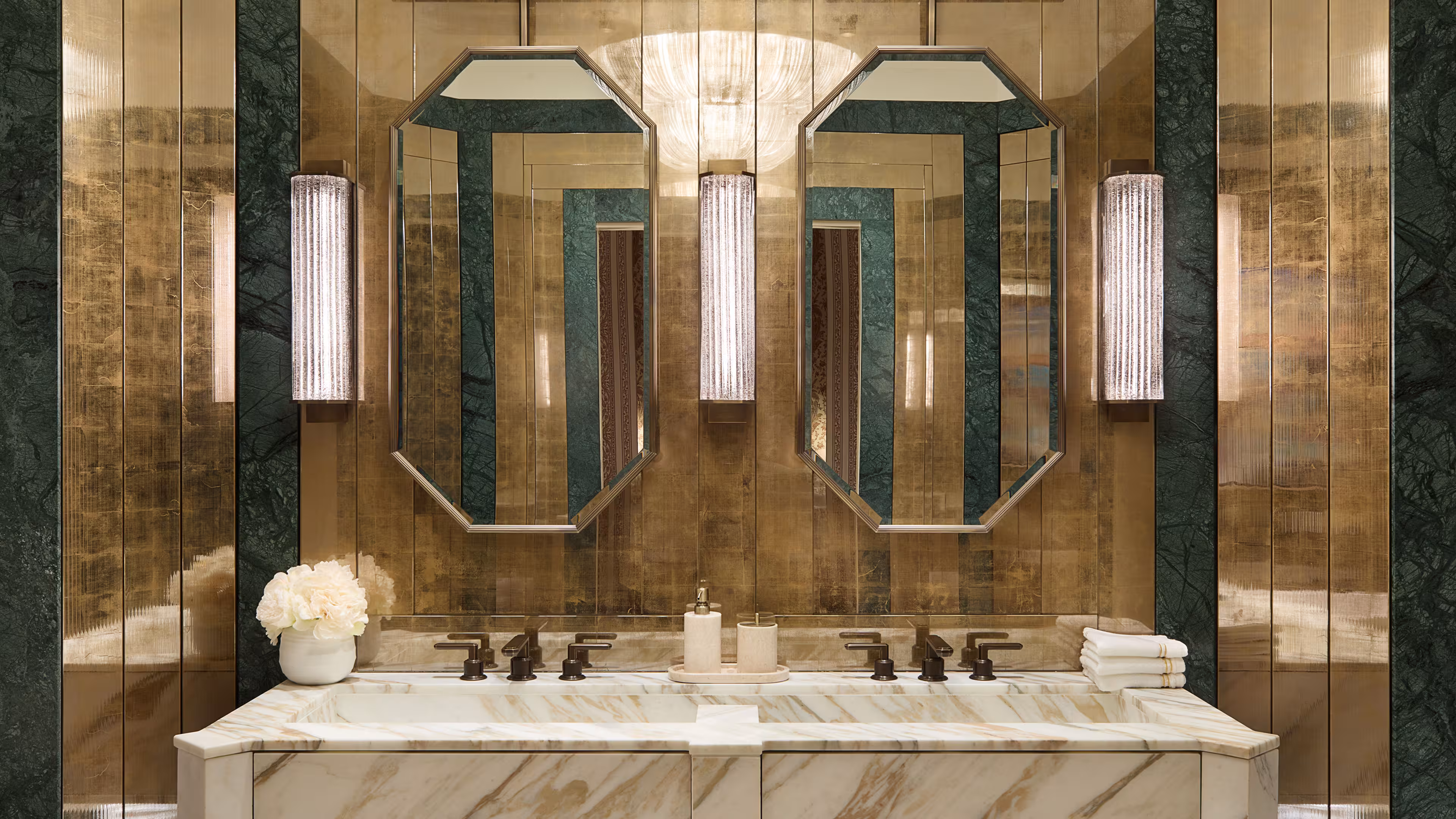 Art Deco bathroom, showing gold paneling, two mirrors and three wall mounted lights. Directly below there’s a large double marble sink with brass hardware.