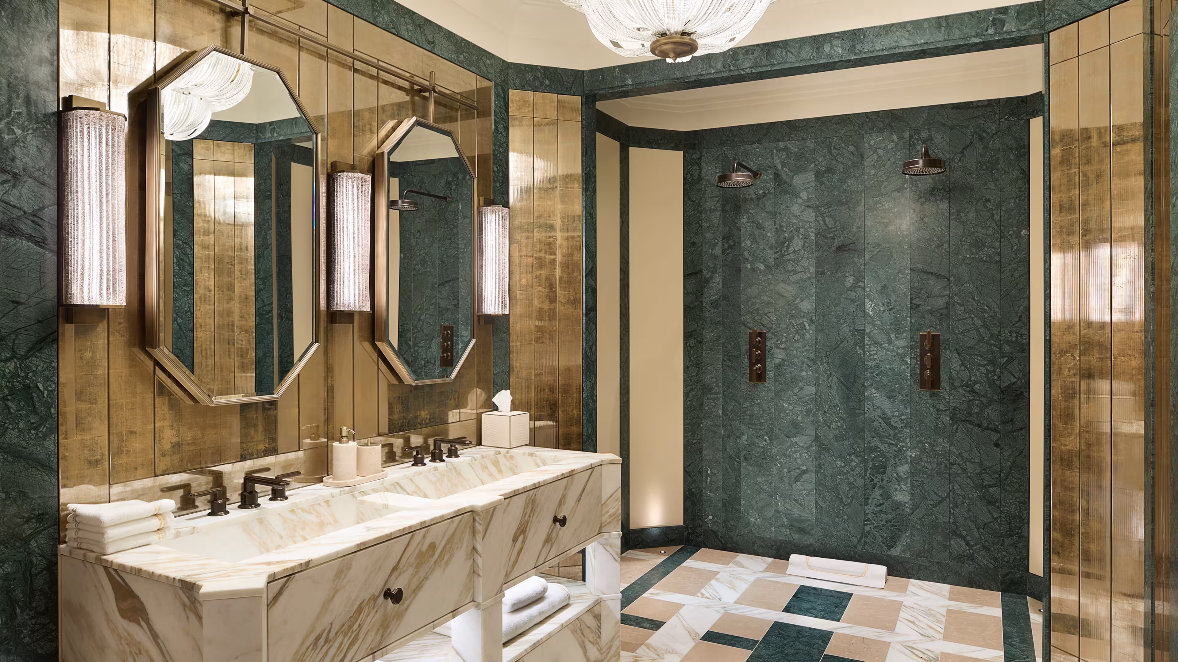 Art Deco bathroom, showing gold paneling, two mirrors and three wall mounted lights. Directly below there’s a large double marble sink with brass hardware to the left. The far wall has a double shower against green marble walls with brass hardware.