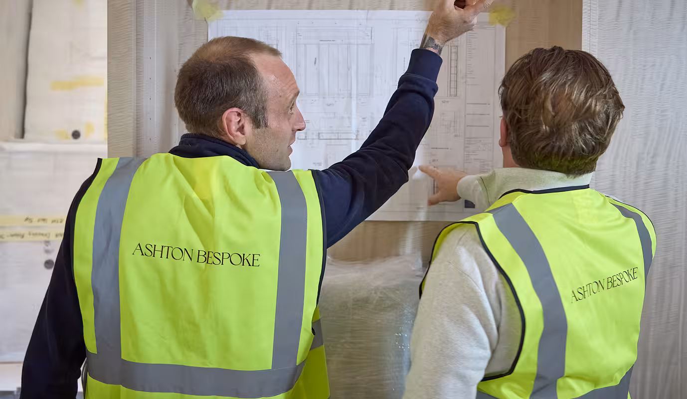 Two workman wearing high visibility tops, with Ashton Bespoke logos on the back. Stood in front of technical drawings on the wall