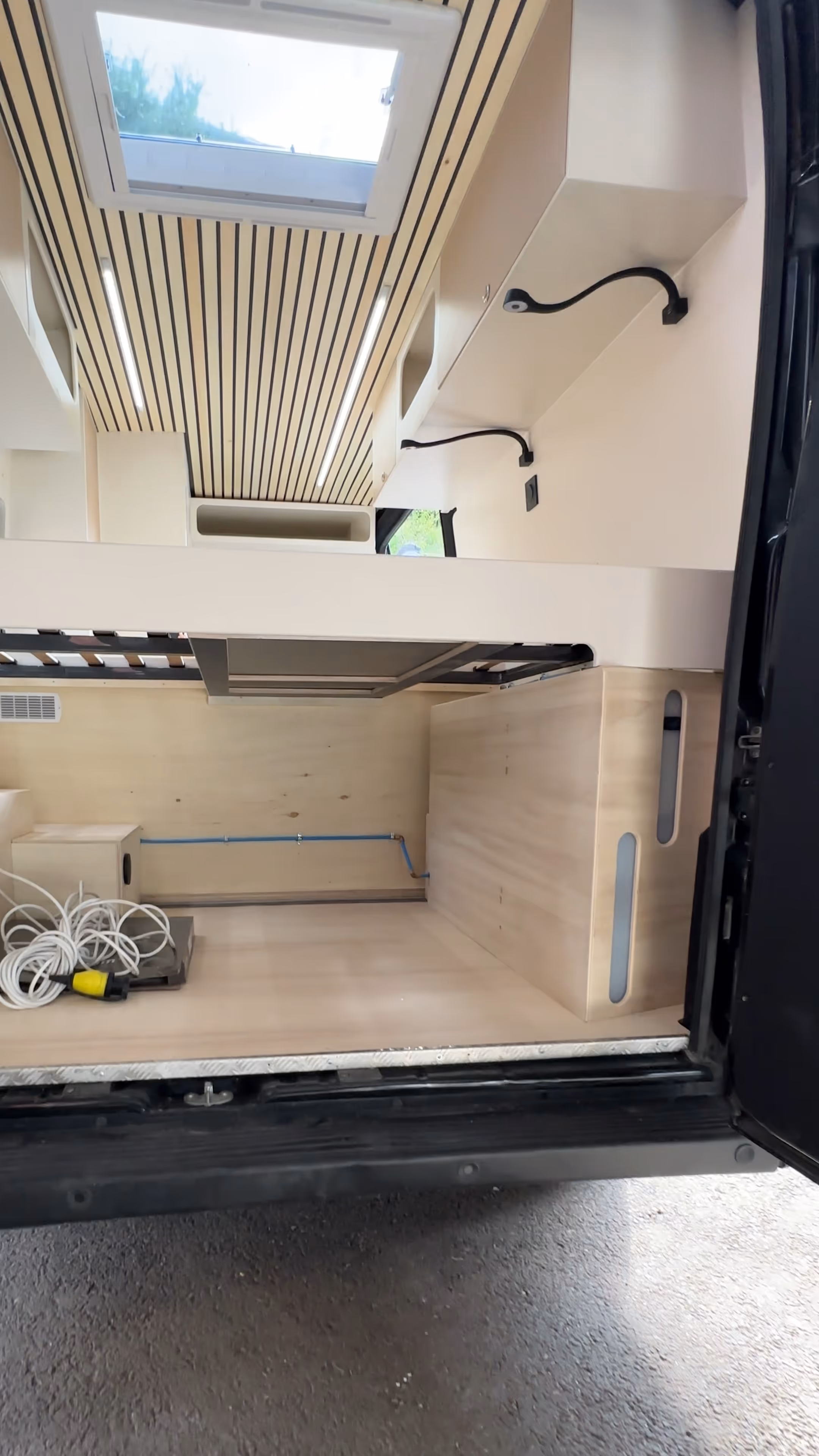 Intérieur d'un van aménagé avec un lit escamotable, un plafond en bois avec un hublot et deux lampes intégrées, des rangements muraux et un câble électrique enroulé sur le sol conçu par Javan Campers