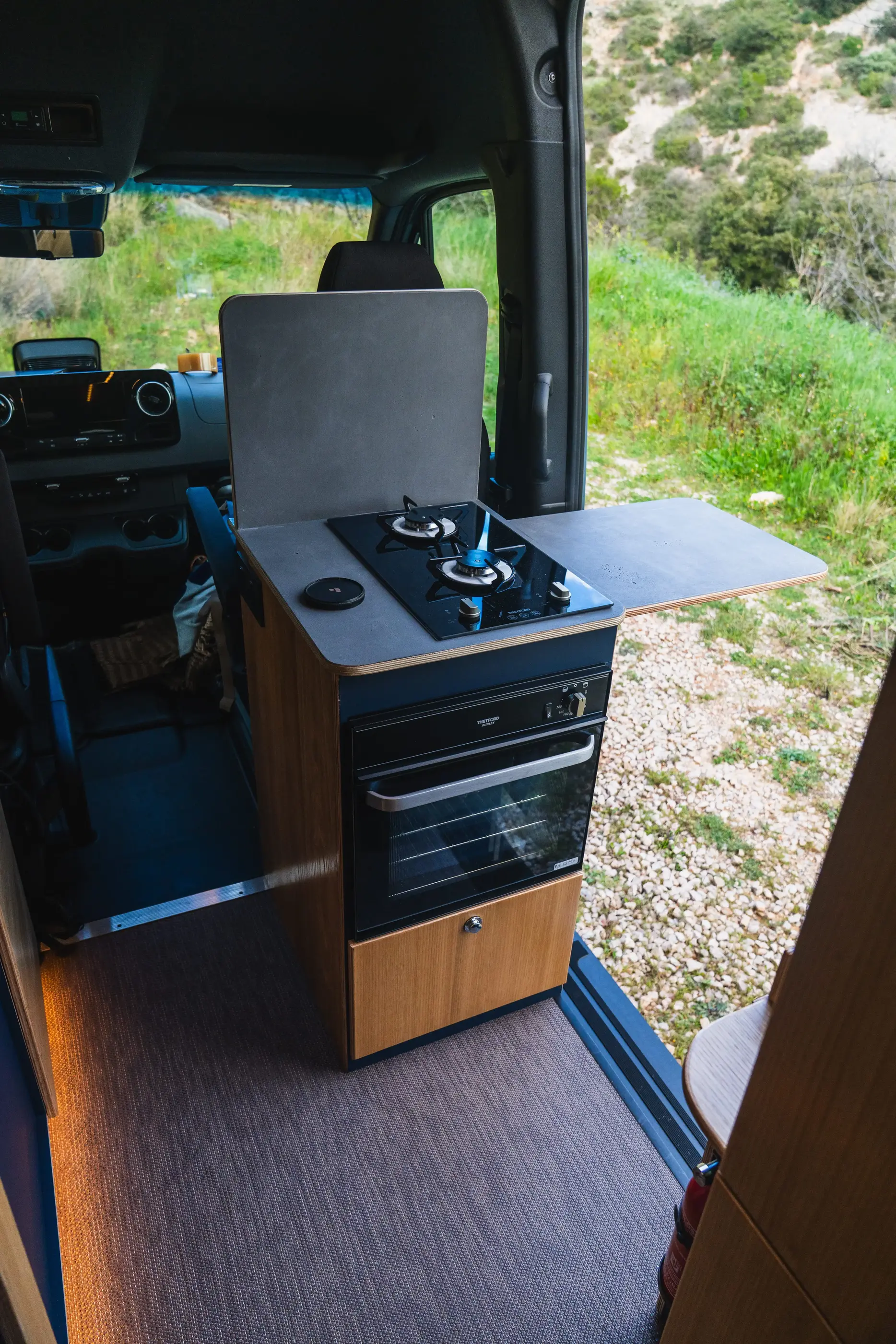 Cuisine compacte avec un réchaud à deux brûleurs et un four dans un van aménagé, porte latérale ouverte sur un paysage verdoyant conçu par Javan Campers