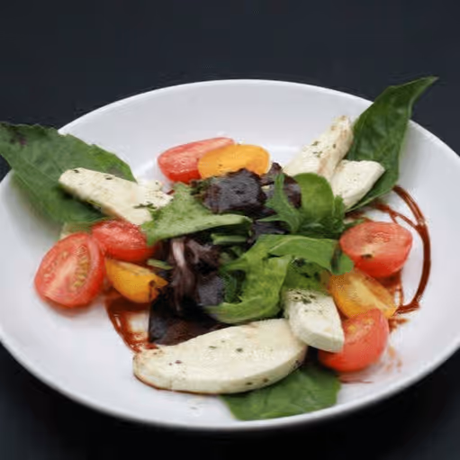 Fresh salad with cherry tomatoes, leafy greens, and mozzarella slices on a white plate. Drizzled with balsamic glaze, the dish is vibrant and appetizing.