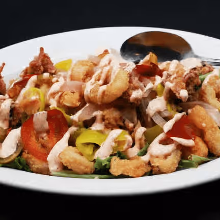 A white plate of fried calamari topped with sliced red and yellow peppers and drizzled with creamy sauce, served with a metal spoon.