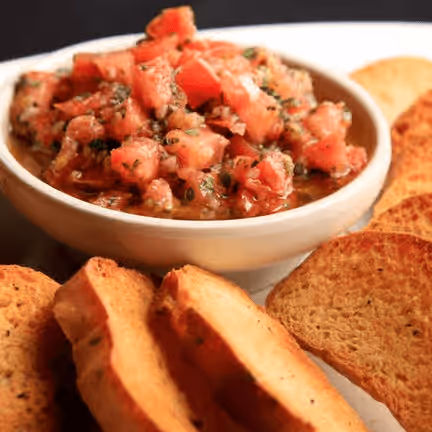 A bowl of vibrant tomato bruschetta, garnished with herbs, sits surrounded by golden, toasted bread slices on a white plate.