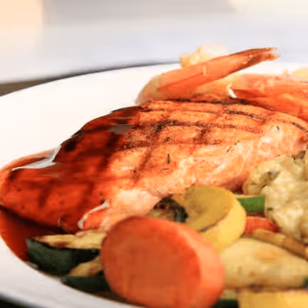 Grilled salmon fillet on a white plate, topped with a glossy sauce. Served with shrimp and roasted vegetables. 