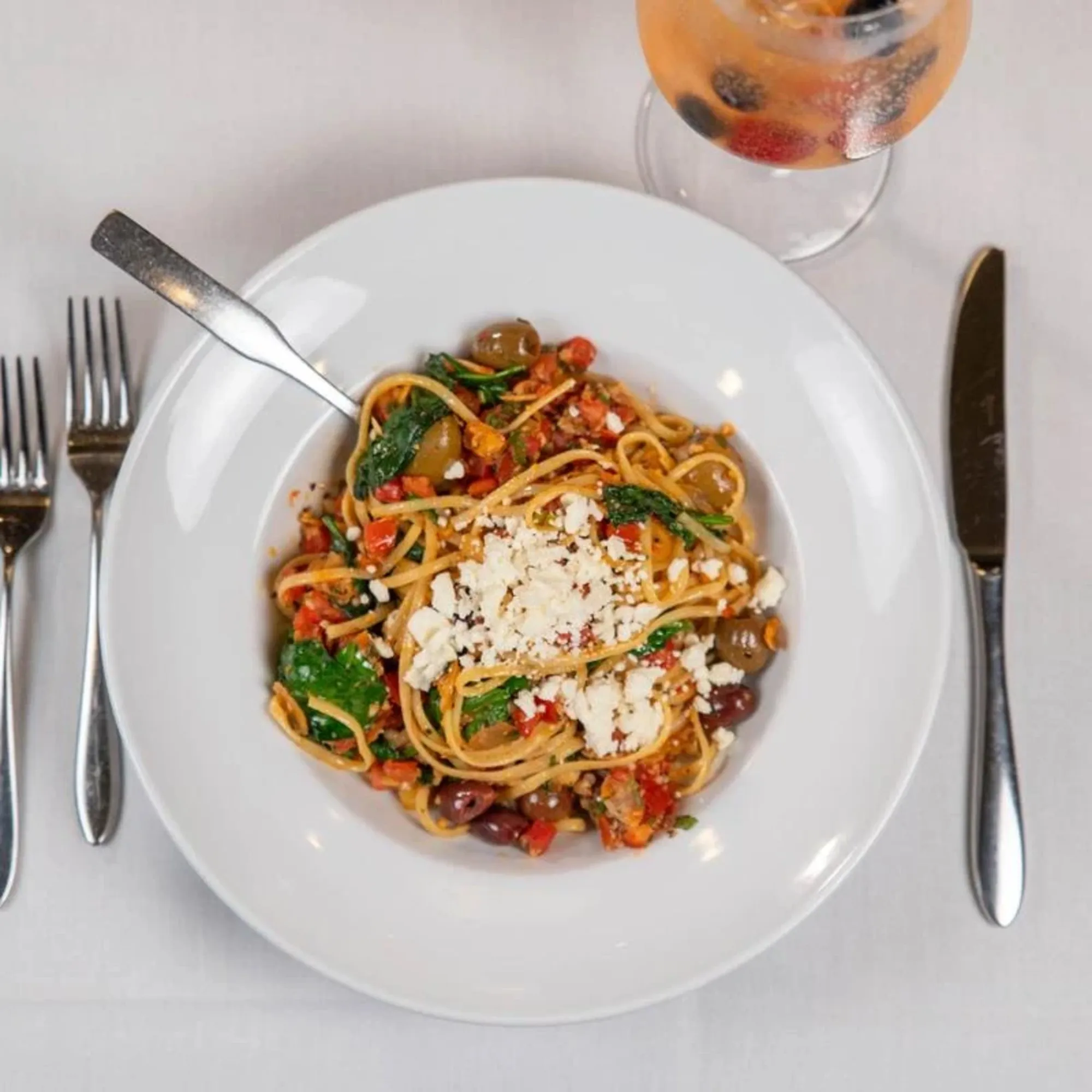 A plate of spaghetti loaded with kale, olives, and tomatoes, topped with crumbled cheese. It's flanked by a fork and knife, with a glass of fruity drink nearby.
