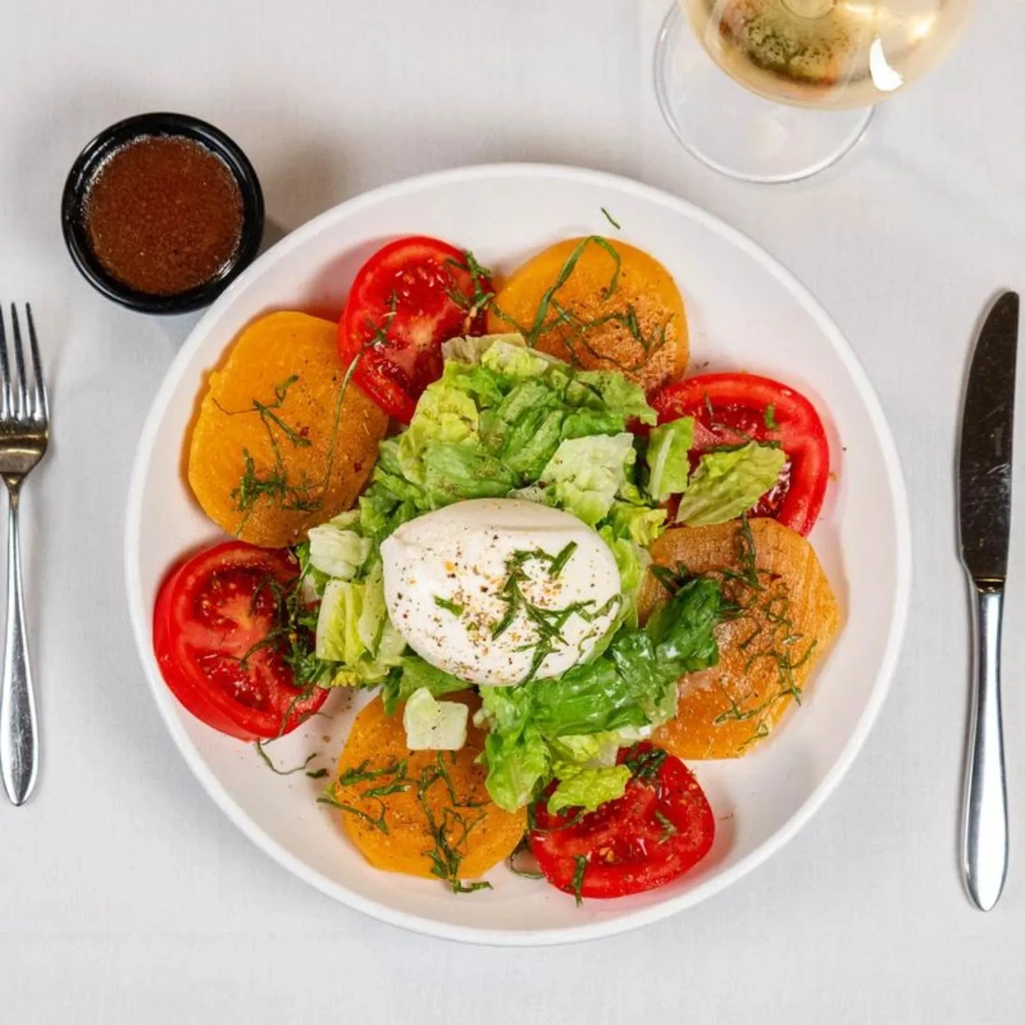 A vibrant salad on a white plate with sliced tomatoes, yellow beets, and leafy greens, topped with burrata. A fork, knife, wine, and dressing bowl surround it.