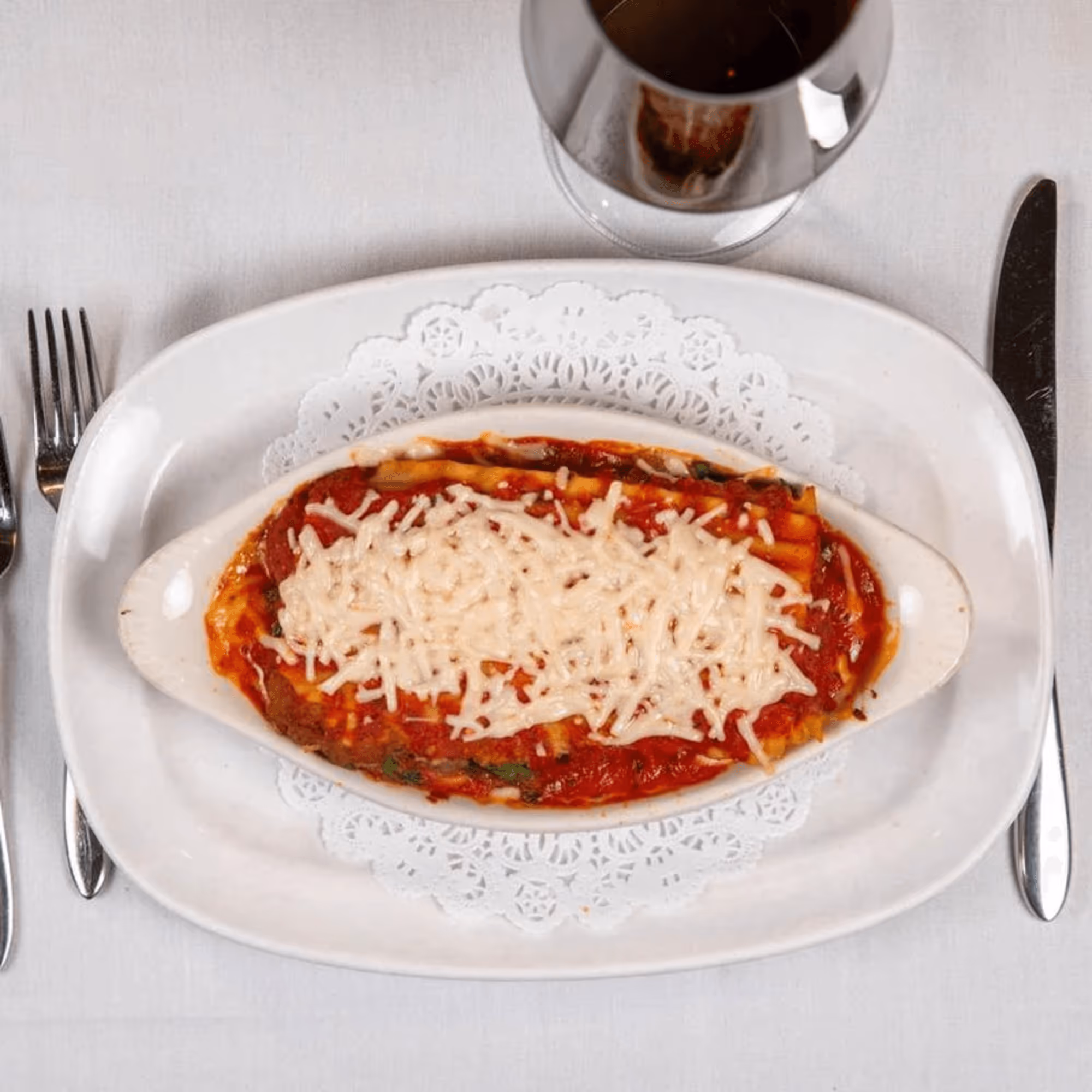 Oval dish of lasagna with tomato sauce and melted cheese, on a white doily-lined plate. Silverware and a glass of red wine are on a tablecloth.