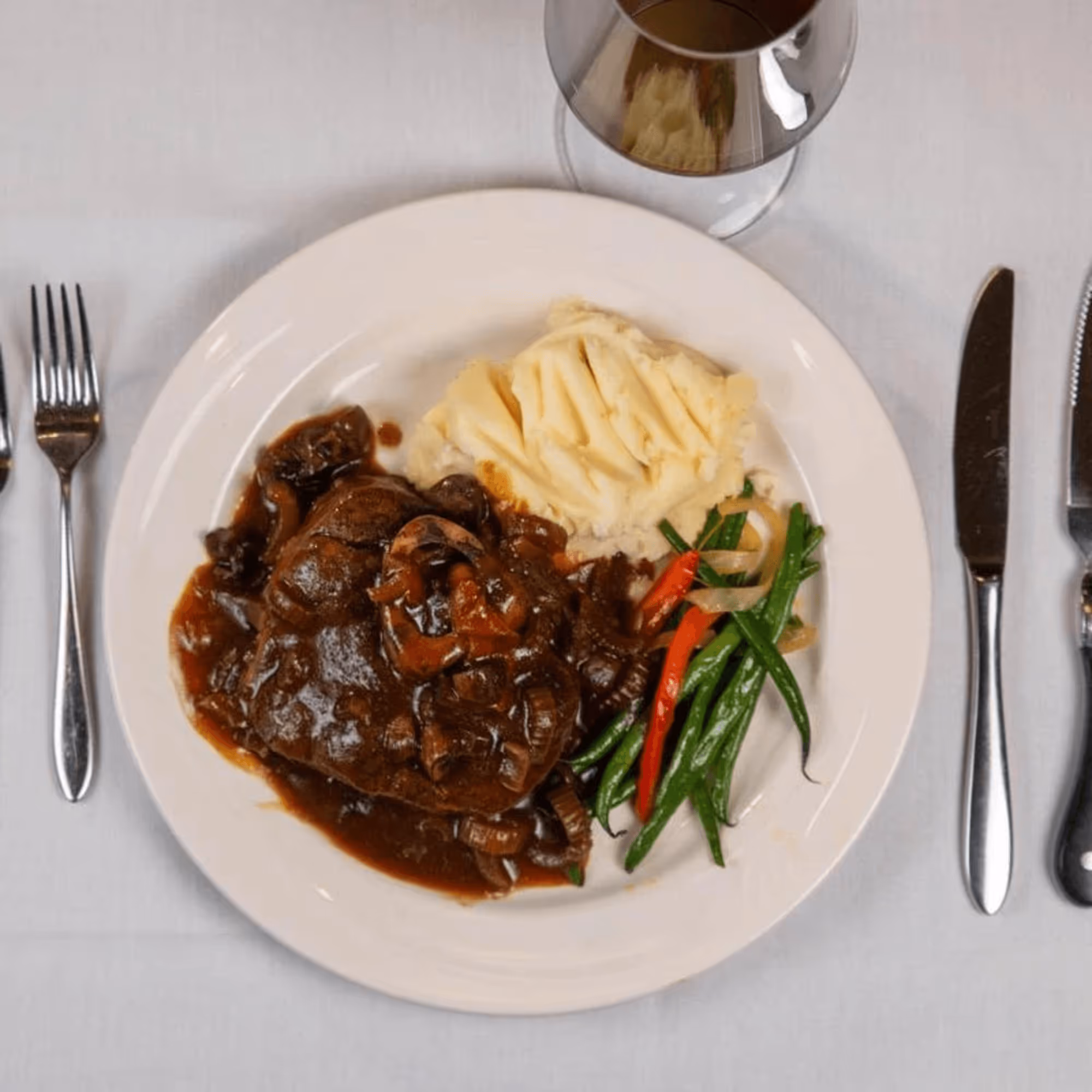 Plate of braised beef in rich sauce, mashed potatoes, and colorful vegetables. Set on white tablecloth with fork and knife, a glass of red wine nearby.