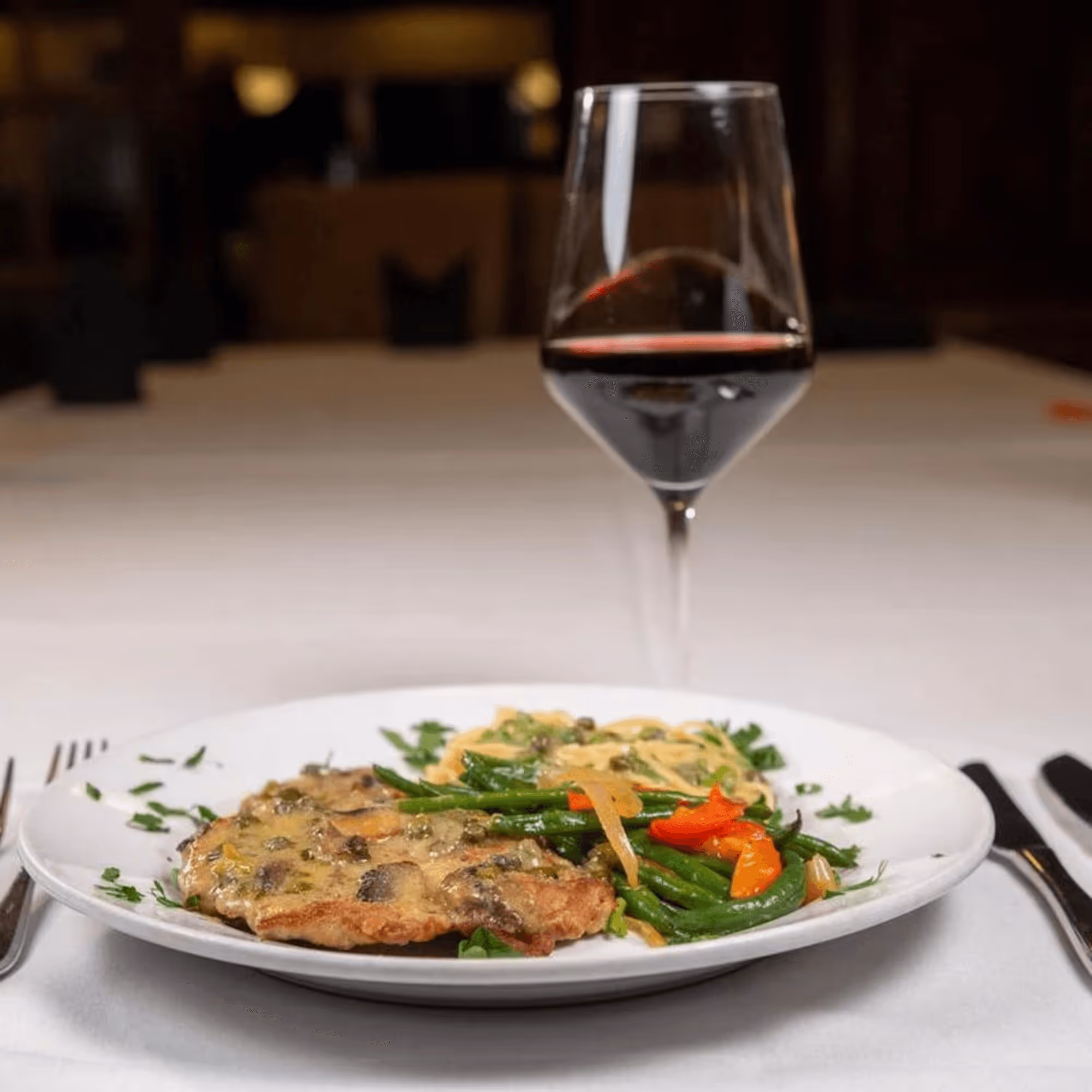 A plate of chicken with a creamy mushroom sauce and sautéed vegetables sits on a white tablecloth, beside a glass of red wine, creating an elegant dining atmosphere.