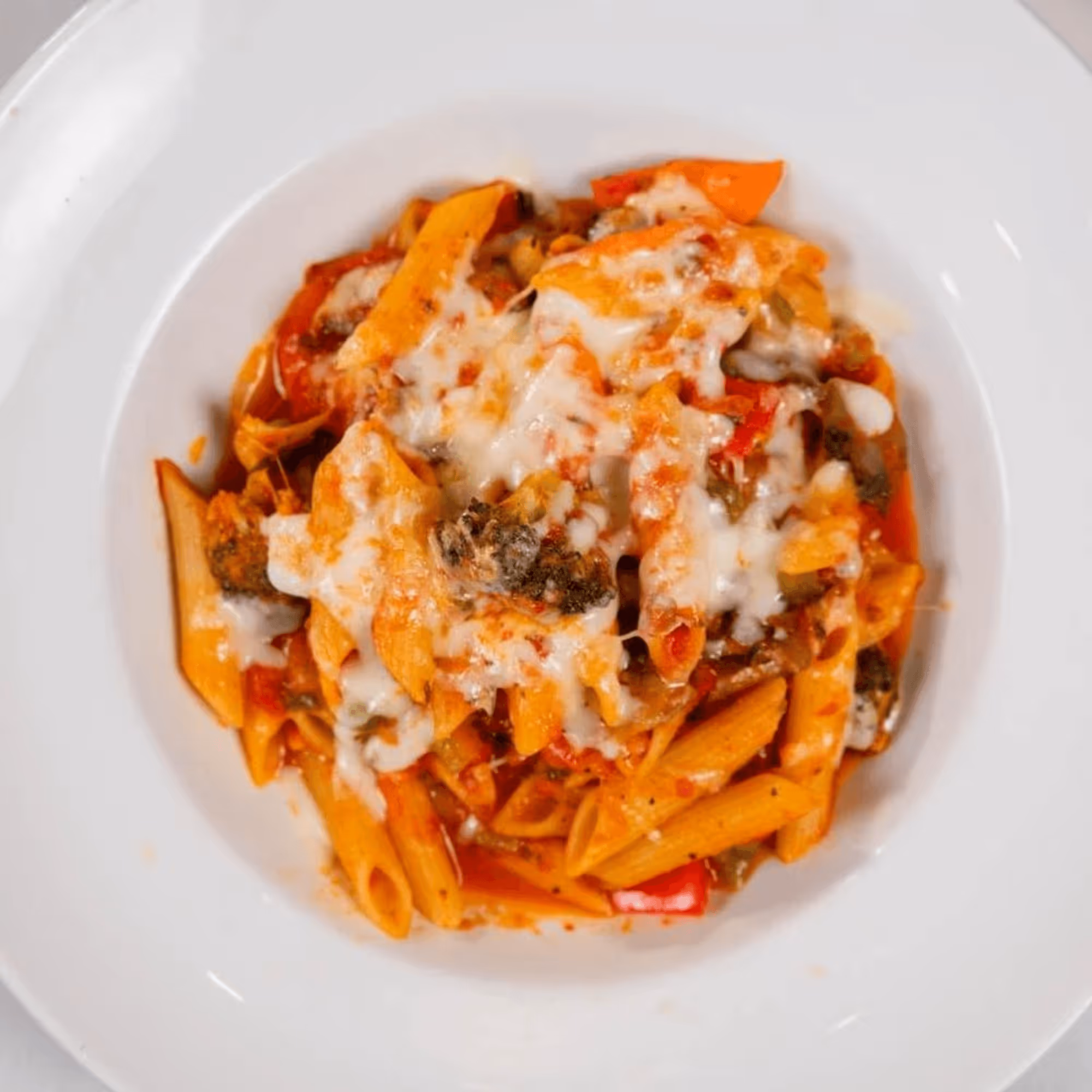 A plate of penne pasta with tomato sauce, topped with melted cheese, red peppers, and mushrooms.