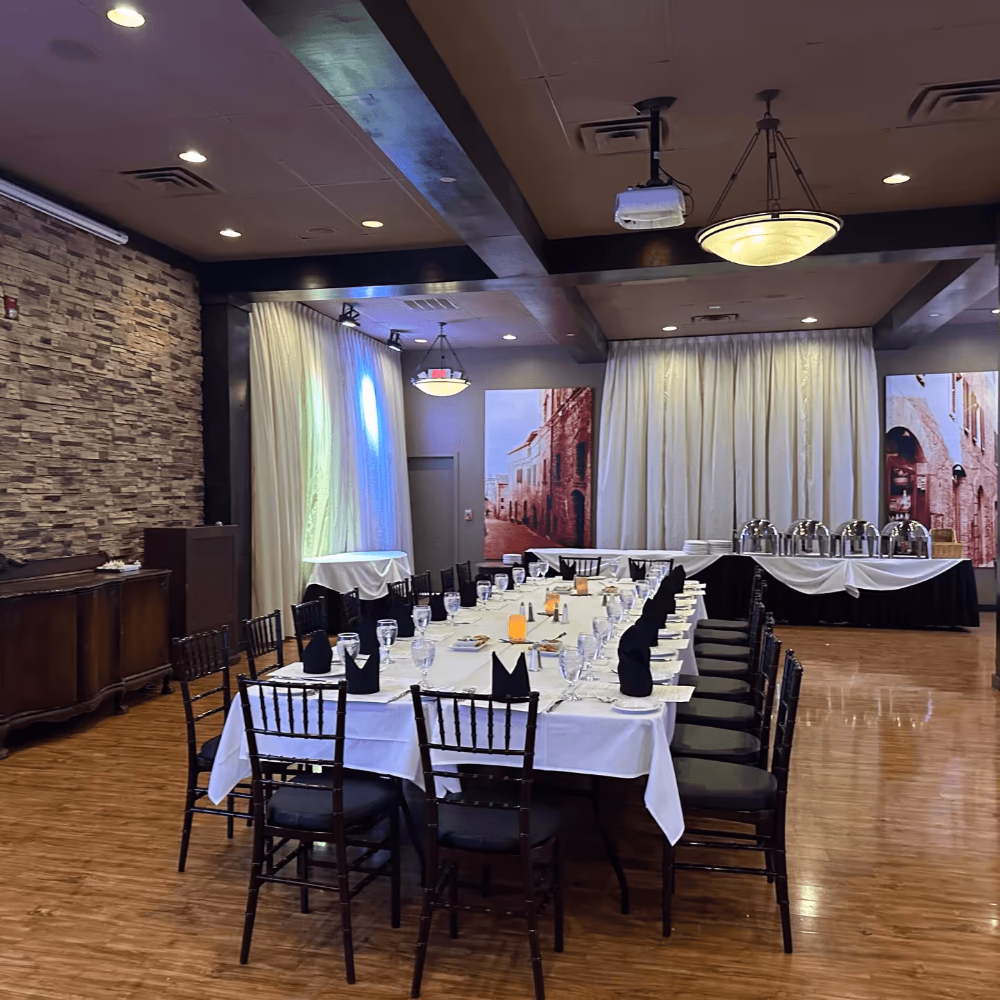Banquet room with a U-shaped table set for a formal event. Table has white tablecloths, glassware, and black napkins. Stone wall and soft lighting create an elegant ambiance.