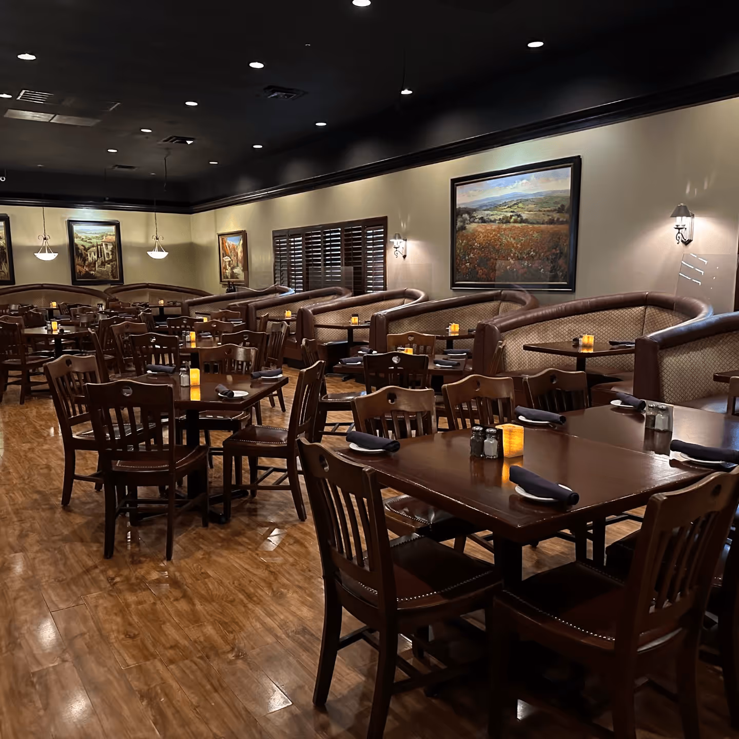 Cozy restaurant interior with dark wood tables and chairs, dim lighting, and warm ambiance. Booths line the walls; paintings and candles add elegance.