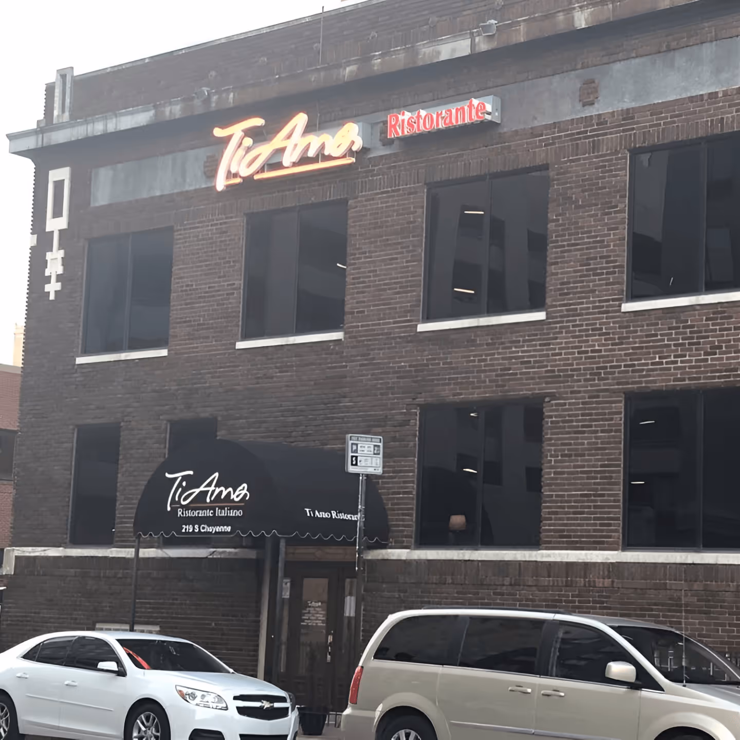 Brick building with neon "Ti Amo Ristorante" sign above. Black awning covers entrance. A white car and beige van are parked on the street, conveying an urban setting.