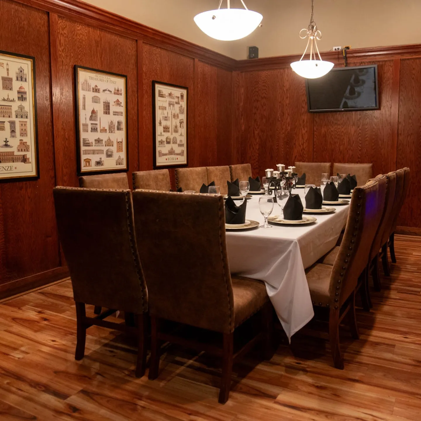 Elegant dining room with a long table set for a formal gathering. Brown cushioned chairs surround the table, adorned with black napkins and glassware. Warm wooden walls and framed art contribute to a cozy, refined ambiance.