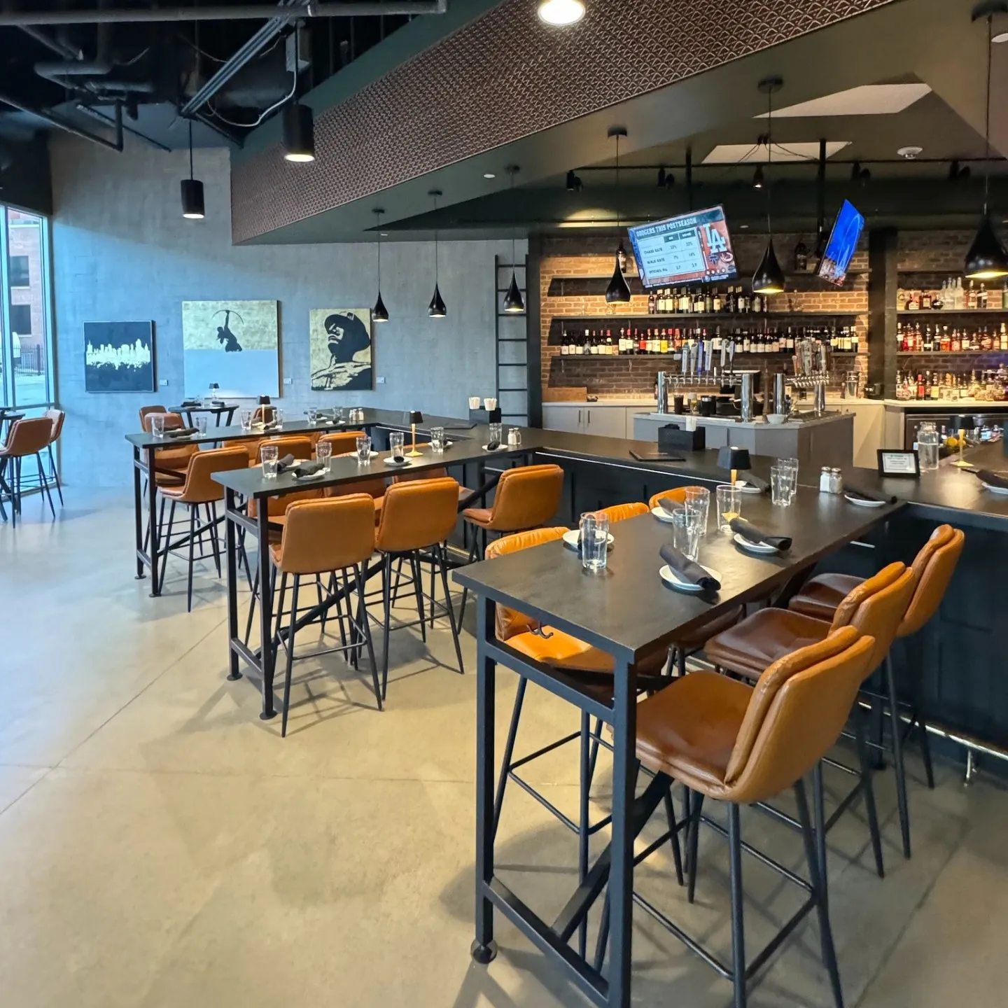 Modern bar interior with high wooden tables and brown leather stools. Hanging lights, shelves with bottles, and wall art create a cozy, stylish ambiance.