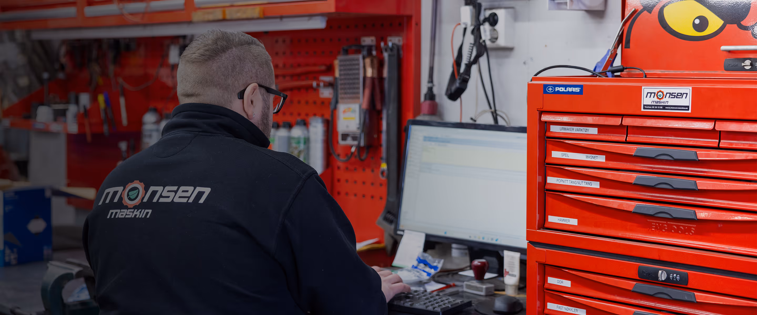 Person med sort jakke med teksten Monsen Maskin jobber ved et skrivebord med dataskjerm og en rød verktøyskuff i verksted.