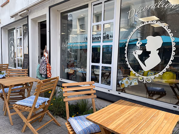 Terrasse avec tables et chaises en bois avec coussins devant une boutique à grande vitrine décorée d'une silhouette tenant une tasse de café.