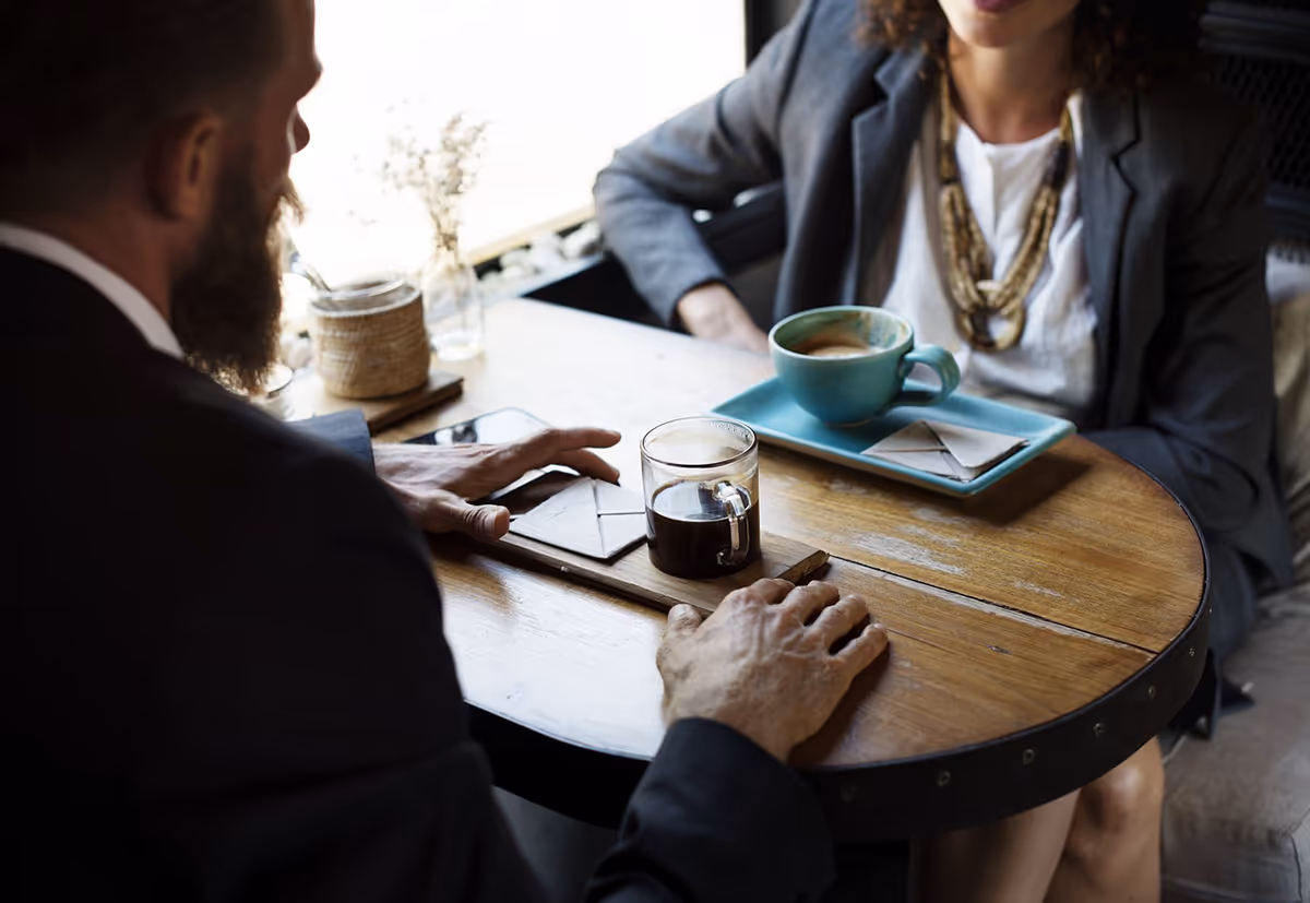 Deux personnes en costume discutant autour d'une petite table ronde avec du café et des enveloppes.