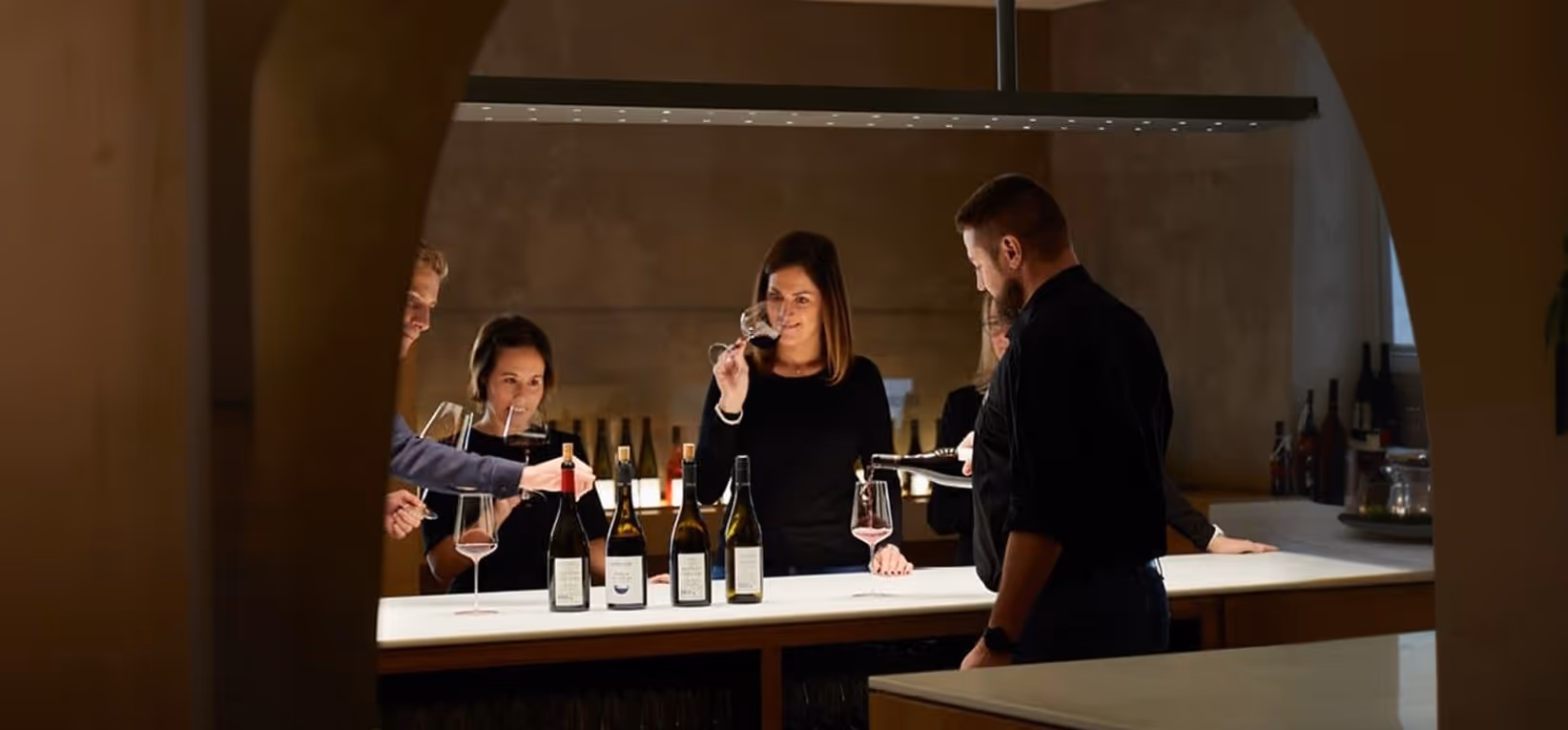 People tasting wine at a bar with several wine bottles on the counter under pendant lighting.