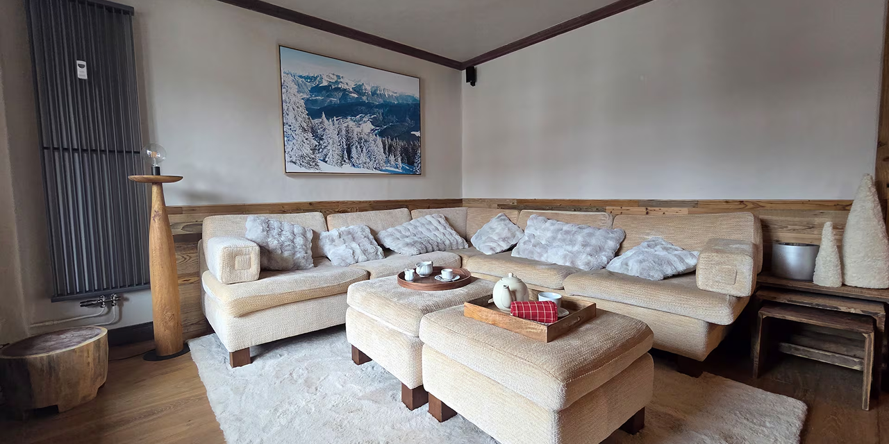 Cozy living room with beige sectional sofa, plush gray cushions, ottomans with tea sets, wooden floor, and snowy mountain landscape painting on the wall.