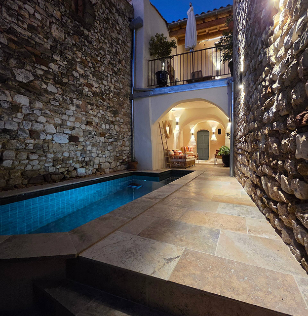 Narrow courtyard with a small blue-tiled pool between stone walls, a tiled floor, and cozy seating under arches illuminated at dusk.