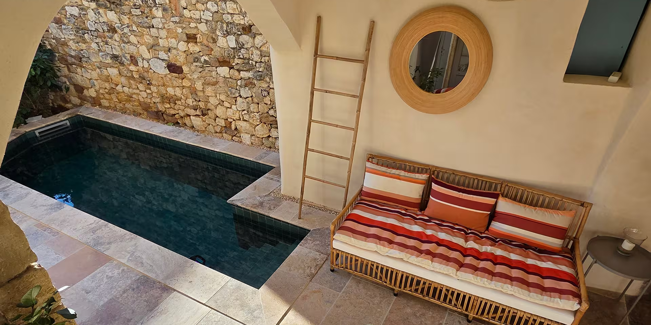 Cozy outdoor area with a small stone pool, striped cushioned wicker sofa, round wooden mirror, and a wooden ladder against a beige wall.