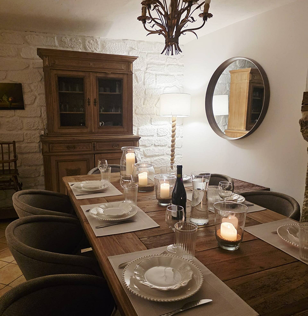 Rustic dining room with a wooden table set for six, featuring white plates, glasses, lit candles, a wine bottle, a tall lamp, and a round mirror on the wall.