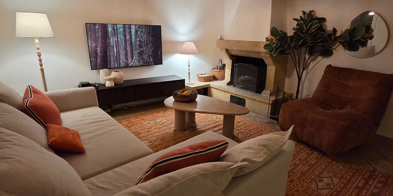 Cozy living room with beige sofa and orange cushions, wooden coffee table, brown corduroy armchair, fireplace, TV mounted on wall, and a tall floor lamp.