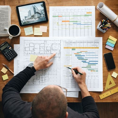 Person reviewing architectural floor plans and project schedules at a wooden desk with a tablet, coffee, calculator, pens, sticky notes, and drafting tools.