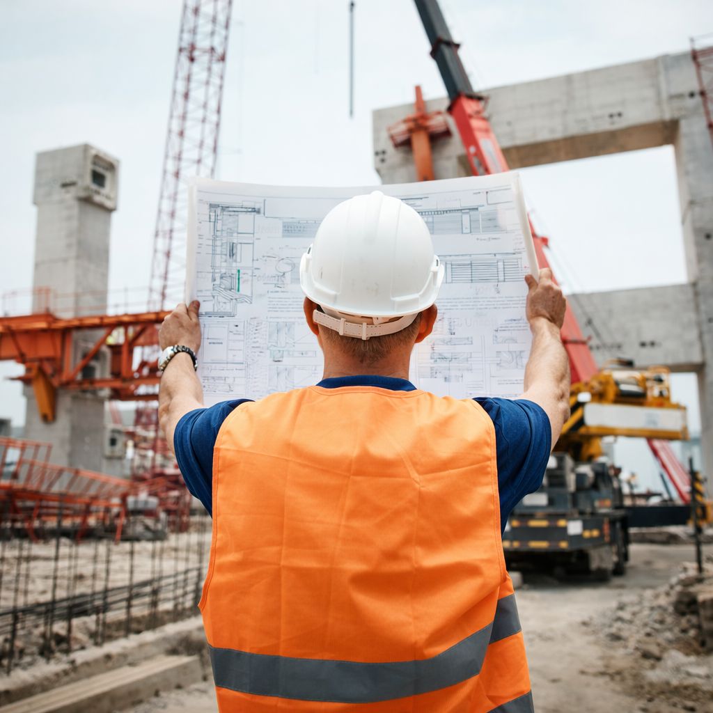 Construction worker in white hard hat and orange safety vest reviewing architectural blueprints at a construction site with cranes and concrete structures.