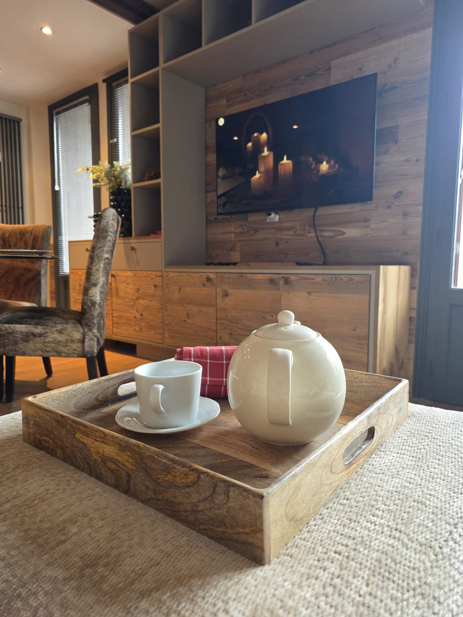 Wooden tray on beige fabric surface holding a white ceramic teapot, a white teacup on a saucer, and a red checkered napkin in a cozy living room.