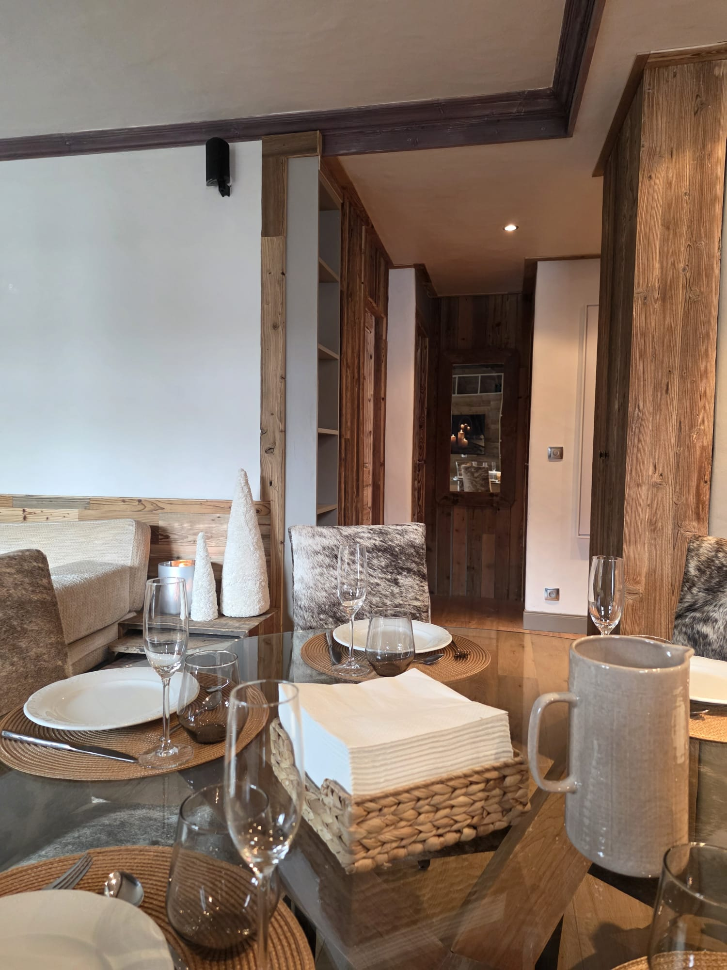 Rustic dining room with a glass table set with white plates, brown placemats, clear glasses, and woven basket of napkins.