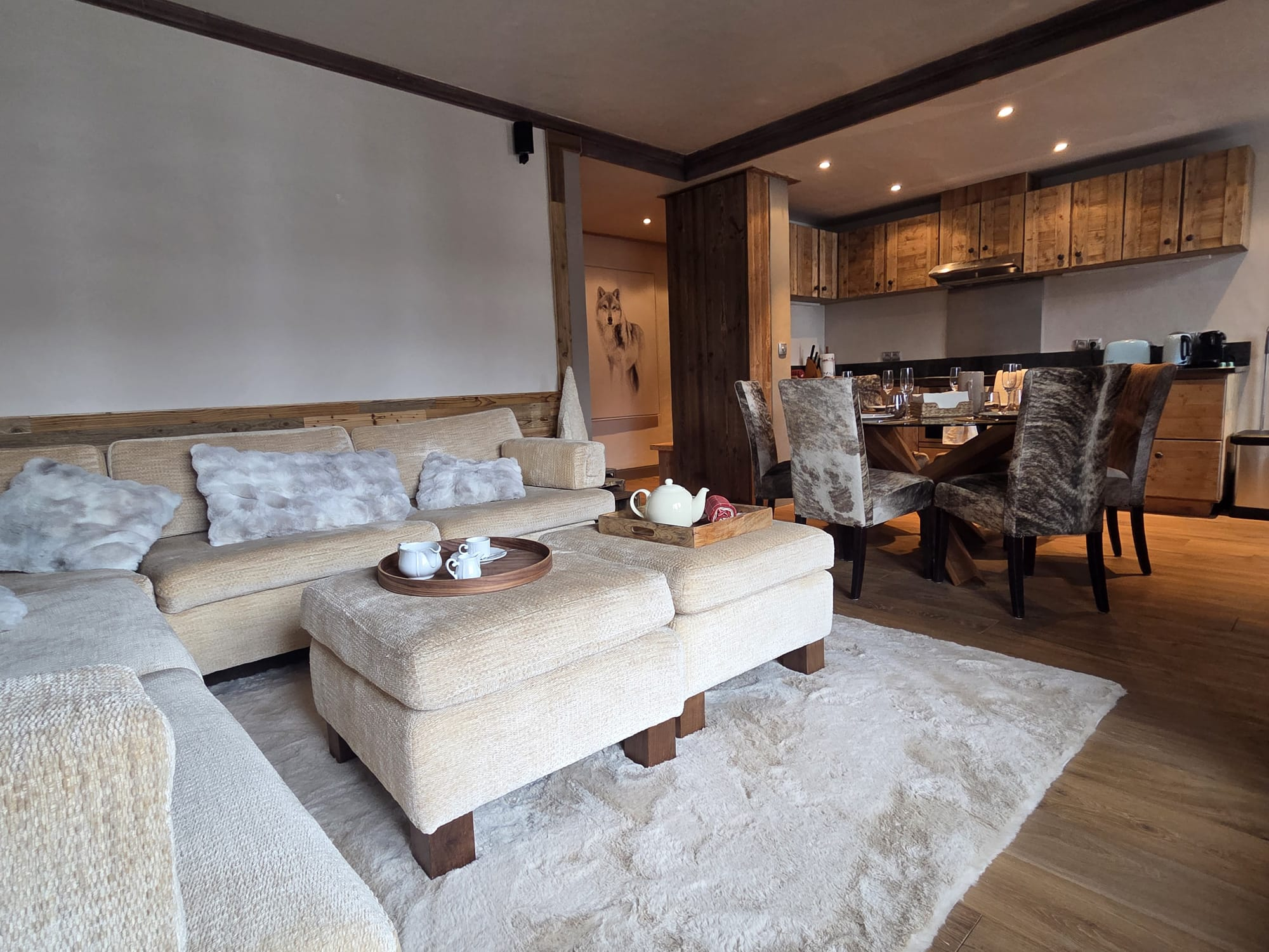 Cozy living room with beige sofas and furry pillows, ottomans with tea set, dining table with patterned chairs, and rustic wooden kitchen cabinetry.