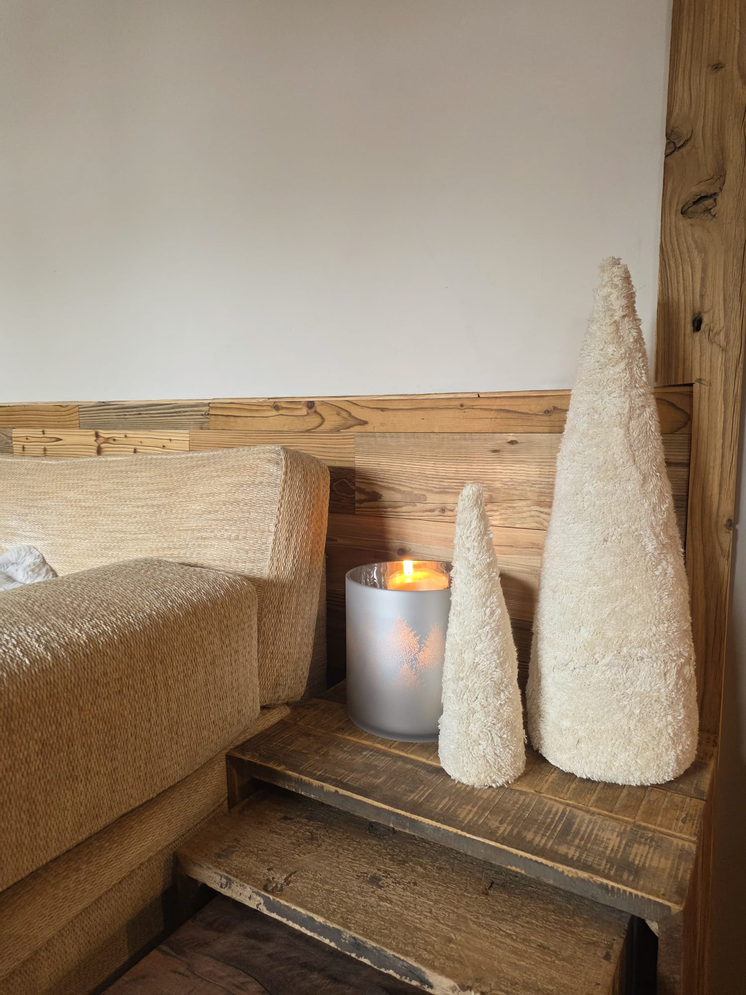 Cozy corner with a beige textured sofa, wooden side table holding a lit candle in a frosted glass holder, and two cream-colored textured cone-shaped decorations.